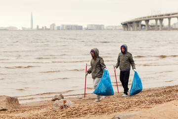 Two environmental activists, young man and woman, walking along sea coast and cleaning beach using trash pickers and plastic bags