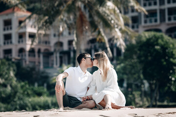 Romantic couple on the beach sitting on the sand. Blurred hotel background. Trip to warm destination