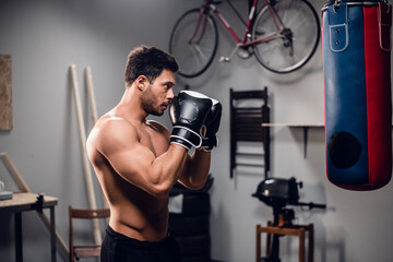 Fototapeta na wymiar a young athlete with a muscular torso is warming up with a punching bag in his garage