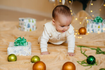 Fototapeta premium Cute smiling baby is lying under a festive Christmas tree and playing with gifts. Christmas and New Year celebrations
