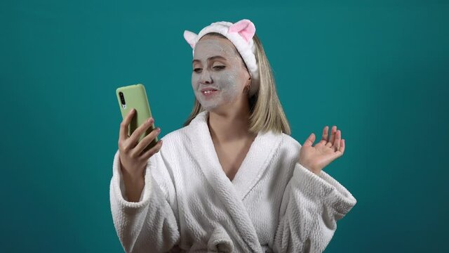 A woman in a white robe, a mask of carbonated bubble clay on her face, talks on a video call
