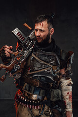 Fototapeta na wymiar Holding shotgun and dressed in dark armour stalker with beard and blood on his face poses in dark background.