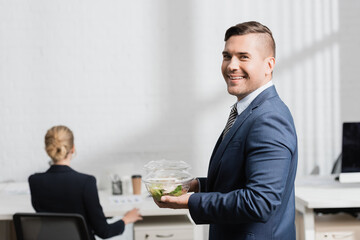  businessman with meal in plastic bowls looking at camera, while standing in office on blurred background