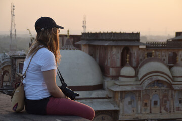 Young woman and solo traveler, sitting on the rooftop watching the sunset over the city skyline with a camera around her neck. Photo taken in India.