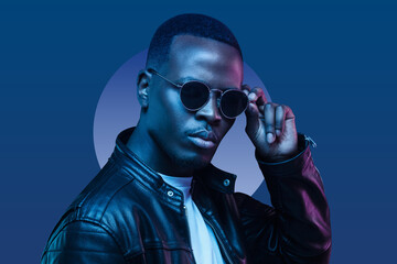 Portrait of african american man, wearing leather jacket, holding sunglasses, isolated on blue background