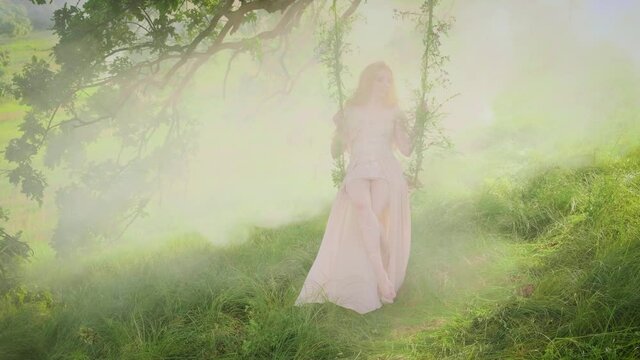 Fantasy woman forest nymph sitting rides on a swing. Summer nature background, white fog smoke, sun rays, divine fairy light, green tree, lawn. Vintage Creative Silk Beige Dress, Blond Long Hair