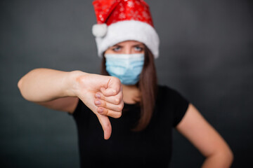 Close-up of thumb down gesturing woman in black T-shirt, medical disposable mask and Christmas cap on gray background.