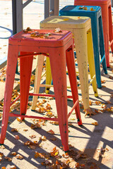 Fototapeta na wymiar Colorful metal stools outside a restaurant on a beautiful fall day with leaves decorating the scene. Red, yellow, orange and turquoise stools.