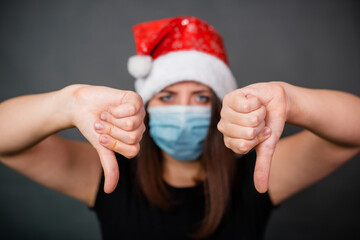 Fototapeta na wymiar Close-up of thumb down gesturing woman in black T-shirt, medical disposable mask and Christmas cap on gray background.