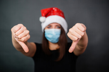 Fototapeta na wymiar Close-up of thumb down gesturing woman in black T-shirt, medical disposable mask and Christmas cap on gray background.