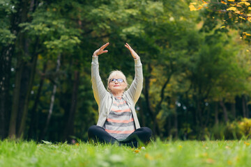 Beautiful senior woman on a walk in the park, performs exercises sitting on the grass, morning in the park