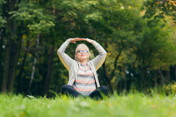 Beautiful senior woman on a walk in the park, performs exercises sitting on the grass, morning in the park