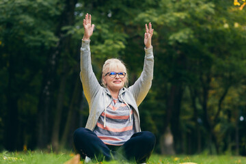 Beautiful senior woman on a walk in the park, performs exercises sitting on the grass, morning in the park