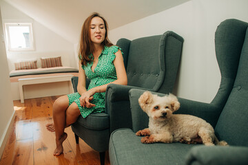 Young Asian pregnant woman in green dress sitting in armchair in bedroom looking at small miniature poodle dog