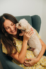 Young pregnant woman in yellow dress holding, petting and playing with miniature poodle dog while sitting in armchair at home 