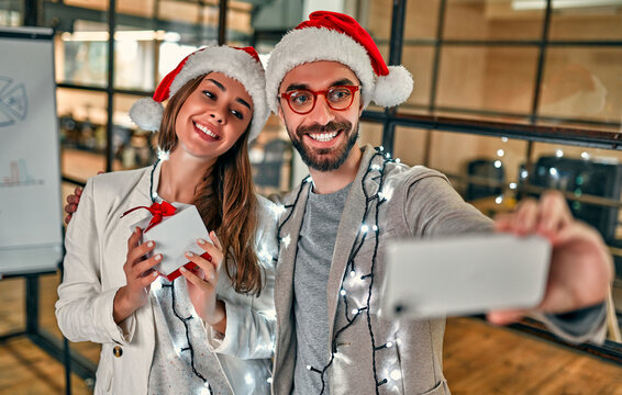 Happy New Year and Merry Christmas! Two young creative people in Santa hats exchange gifts and take smartphone selfies on their last working day.