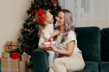 Obraz na płótnie Canvas Happy mother and child girl with gift box at home near the Christmas tree. Merry Christmas and Happy Holidays. Happy New Year 2021. Mother hugging cute baby girl together at home. 