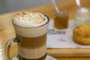 Café capuchino servido en taza de vidrio con crema y canela, tabla con escones y dulce cacero