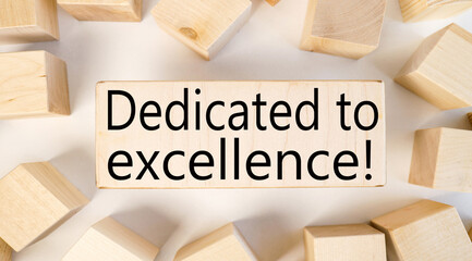 DEDICATED TO EXCELLENCE, text on a block of wood on a light background