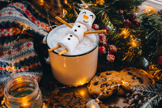 Christmas food white cup with hot chocolate snowman marshmallows. homemade cookie, cinnamon sticks fir xmas tree branches with warm garland lights. New year celebration. Holiday family fun in winter.