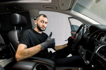 Young Caucasian male car wash detailing worker using cleaning brush and removing dust from car control panel and steering wheel, showing his thumb up and smiling at camera