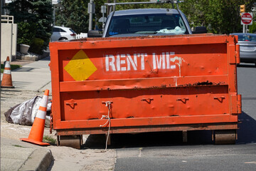 The end of an orange dumpster seen on a street in front of a truck on a sunny day