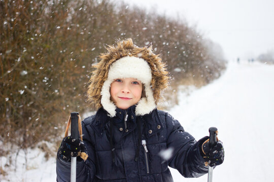 Joyful happy Russian boy wearing traditional ushanka hat and warm outerwear is skiing in winter during snowfall with ski poles on forest road. Winter activities for kids and family. 