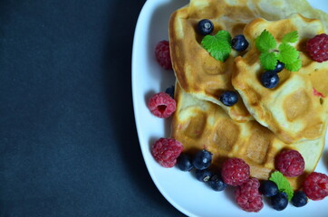 Homemade Belgium Waffles with fresh blueberries for breakfast. White plate, Viennese waffles with raspberries and mint. quick delicious breakfast on black background. top view copy space