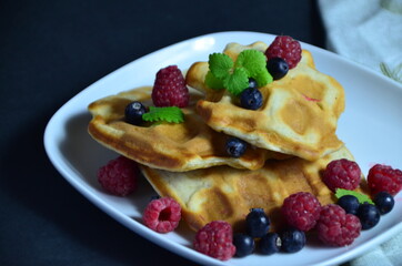 Homemade Belgium Waffles with fresh blueberries for breakfast. White plate, Viennese waffles with raspberries and mint. quick delicious breakfast on black background. top view copy space