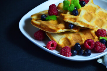 Homemade Belgium Waffles with fresh blueberries for breakfast. White plate, Viennese waffles with raspberries and mint. quick delicious breakfast on black background. top view copy space