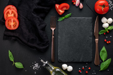 Food background for Italian cuisine. Cooking ingredients for caprese salad with fork and knife around blank slate for recipes, top view, frame, mock up