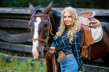 Fototapeta premium Smilng pretty young cowgirl. A cowboy style female in a plaid shirt standing next to the horse and holds the horse by the bridle.