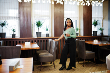 Portrait of attractive young african american woman wearing in green sweater and black jeans pose at restaurant with glass of red wine at hand.
