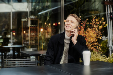 A man is sitting at a table in a cafe on the street Holding a Cup of coffee and talking on the phone.