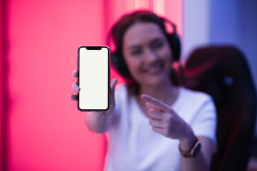 Smiling gamer girl wearing headset showing smart phone and pointing at blank screen with her finger.