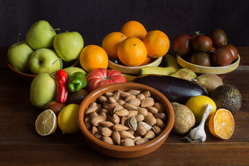 Still life background with whole almonds, garlic, banana, oranges, peppers, avocado, tomato, lemon, pears, and apples on natural dark wood and clay bowls. Celebrations and healthy food.