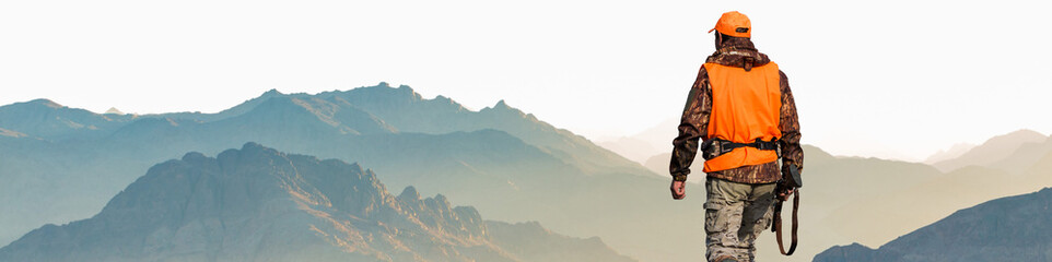 A hunter in camouflage and a gun in his hands in the early morning on top of a mountain stands looking for a target.