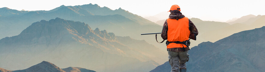 A hunter in camouflage and a gun in his hands in the early morning on top of a mountain stands...