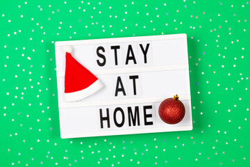 Light box with text STAY AT HOME and Christmas decoration on green background with gold glitter stars. Top view