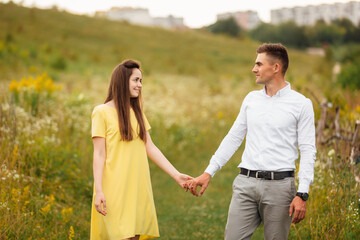 Fototapeta na wymiar young happy couple in love holding hands in a grass field in sunny day. girl in a yellow dress and a guy with a stylish haircut. portrait of couple walking on countryside