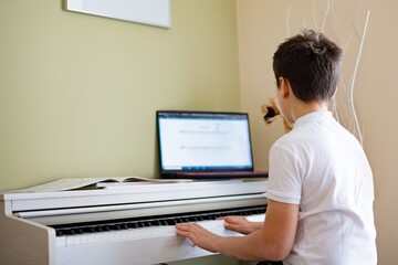 A young boy plays the digital piano with sheet music from his laptop.