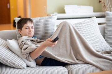 Cute kid is watching TV is lying under a blanket on the sofa with the TV remote in hand.