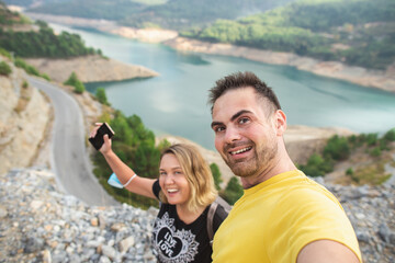 A couple in love takes selfie photo portrait among mountains at Dim Cay reservour, Alanya. Happy hipster students take photo for their blog or social media. Medical mask on the hand of woman..
