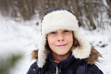 Winter portrait of young joyful happy local Russian boy wearing traditional ushanka hat made of artificial fur and warm outerwear during winter activities outside