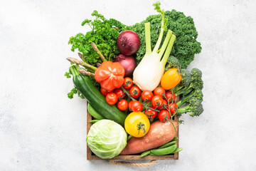 Variety of organic vegetables in brown box, broccoli cauliflower cabbage kale pak choy onions tomatoes. Healthy raw produce on white wooden table, top view, selective focus
