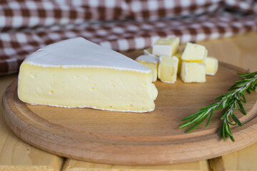A piece of brie cheese on a wooden board, next to sprigs of rosemary. In the background is a kitchen towel. Shallow depth of field