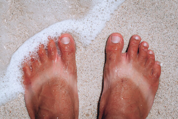 suntanned man feet with stripes of white skin from sandals in summer standing on the sand and sea waves