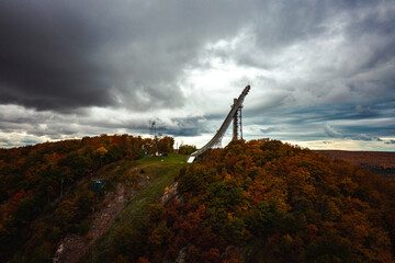 Beautiful view of Copper Peak ski jump and hill sticking up high into the cloudy and overcast sky above surrounded by colorful red, orange, yellow and green tree foliage.