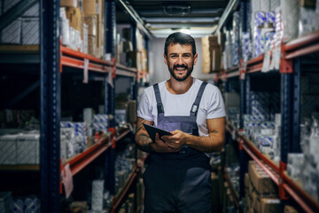 Fototapeta na wymiar Smiling bearded tattooed hardworking employee holding tablet and standing in storage.