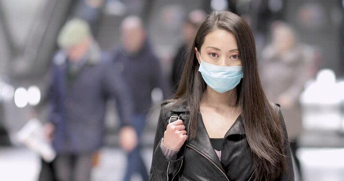 Chinese woman at train station in London wearing face mask to protect from covid 19 coronavirus - young asian woman portrait in a busy public place protecting from virus outbreak - health lifestyle